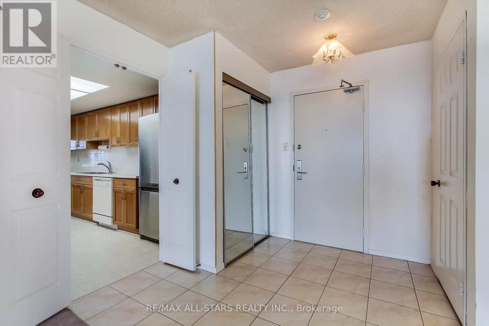 A white door opens to a kitchen with wooden cabinets and a stainless steel refrigerator.
