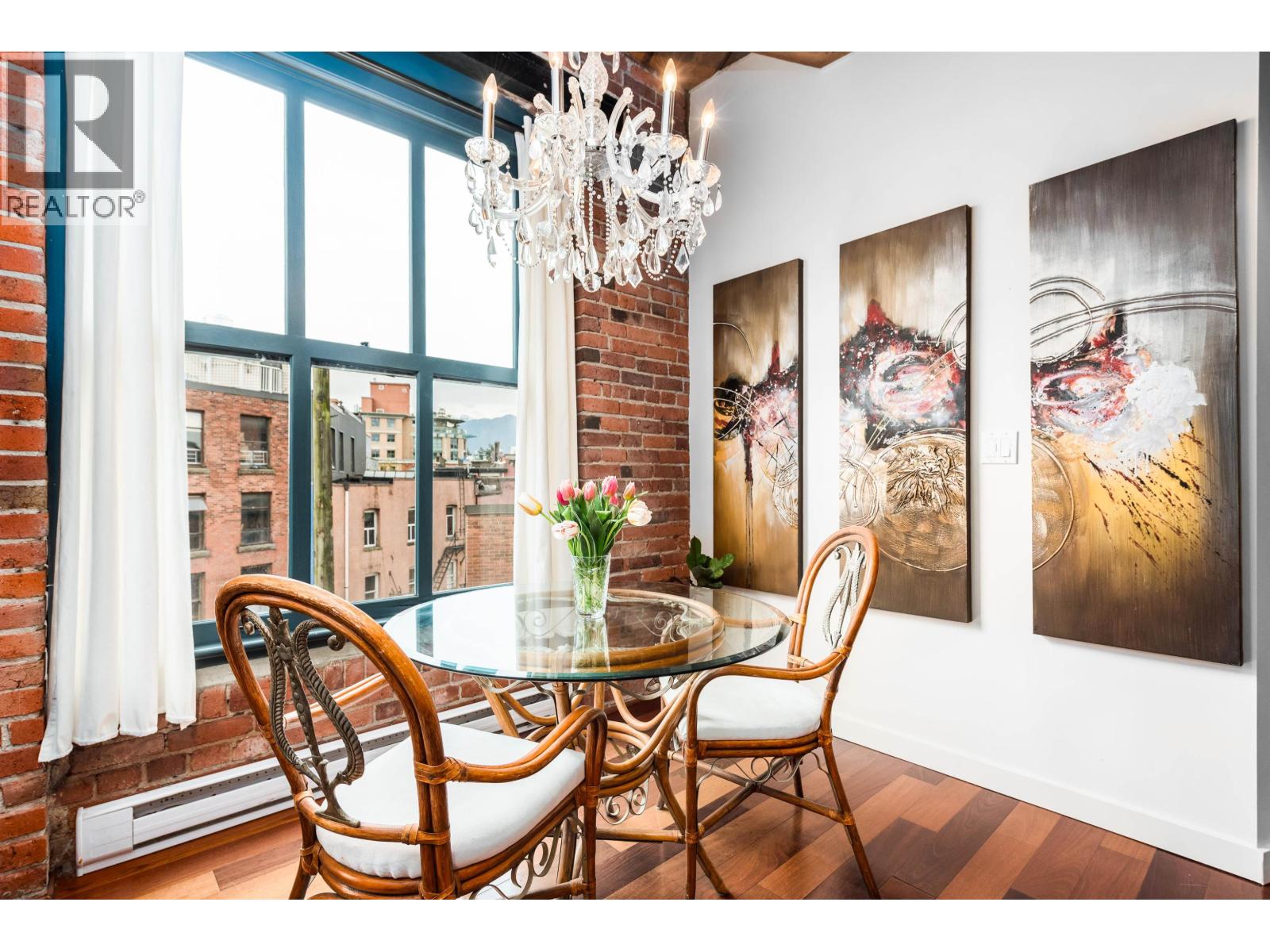 A dining room with a glass table and chairs, a chandelier, and three paintings on the wall.