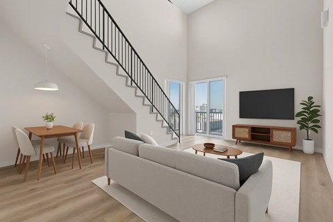 A living room with a grey couch, a wooden dining table with chairs, a staircase with a metal railing, a flat screen TV, and a potted plant.