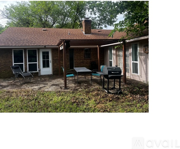 A patio with a grill and chairs is surrounded by a brick house and trees.