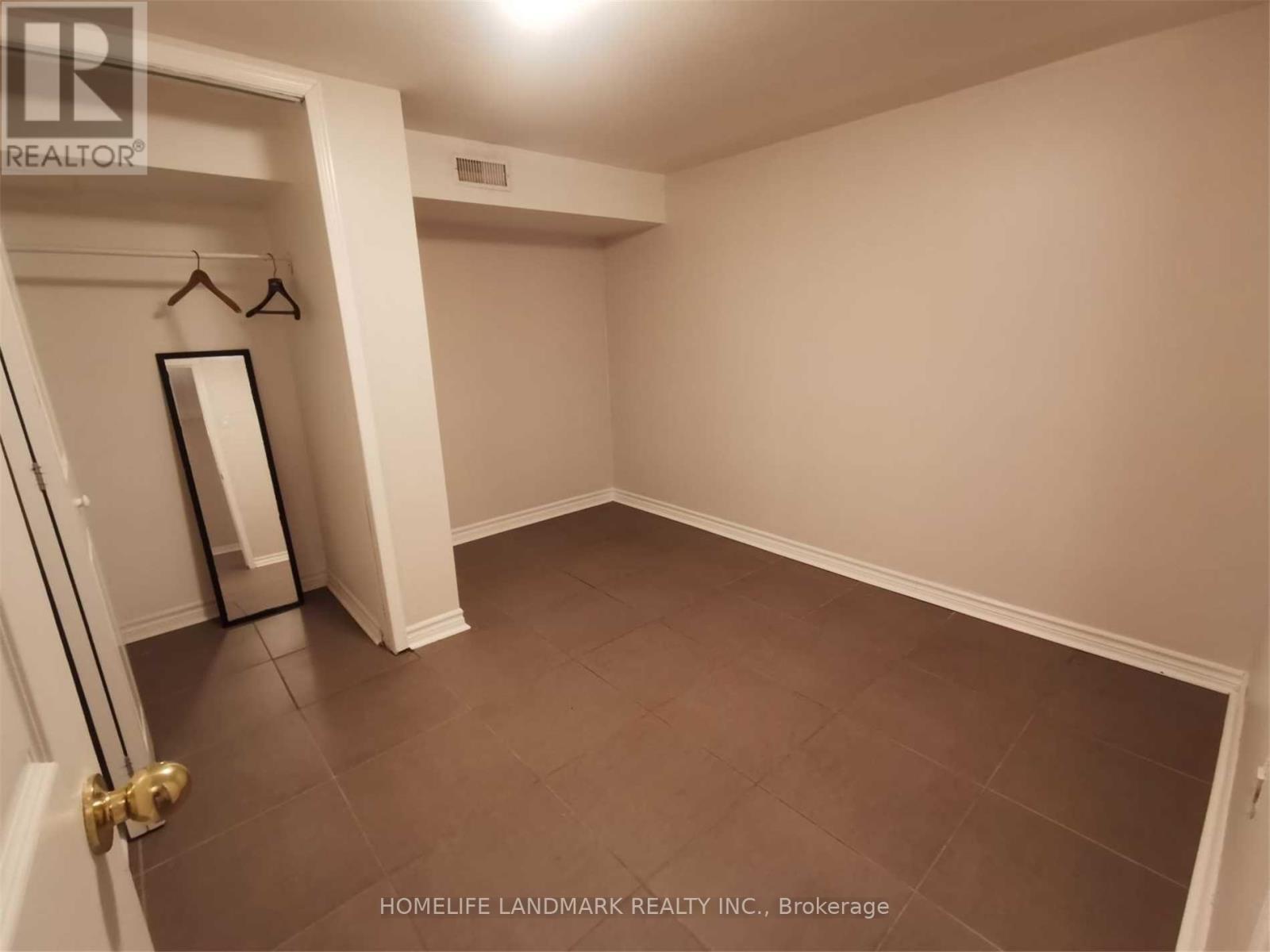 A room with a brown floor and white walls, with a door on the left and a coat rack with a mirror on the wall.