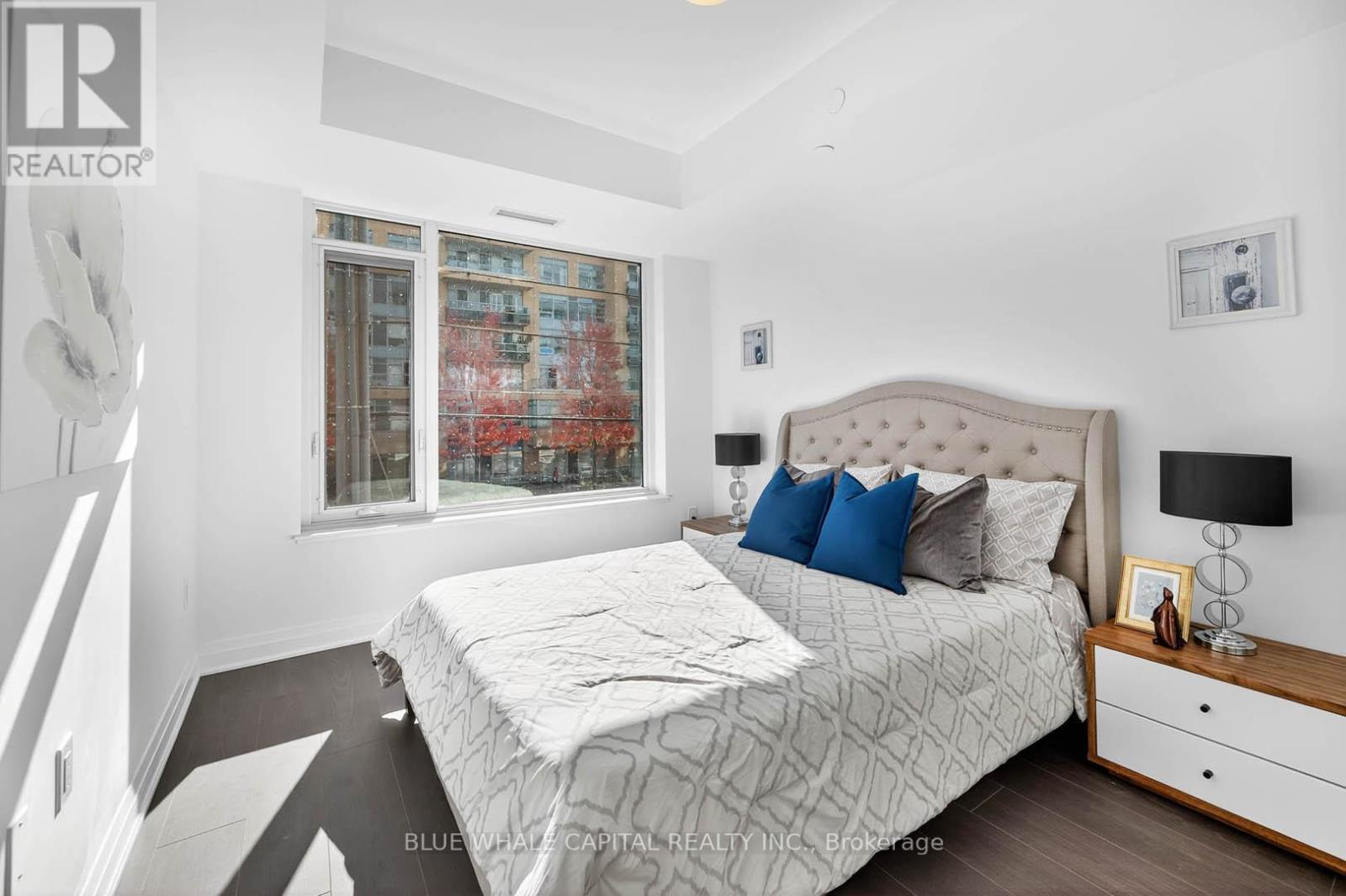 A bedroom with a large bed, a window with a view of a building, and a wooden nightstand with a lamp.