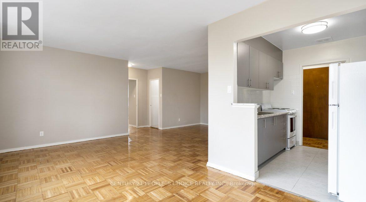 A large empty room with wooden flooring and a kitchen area in the background.