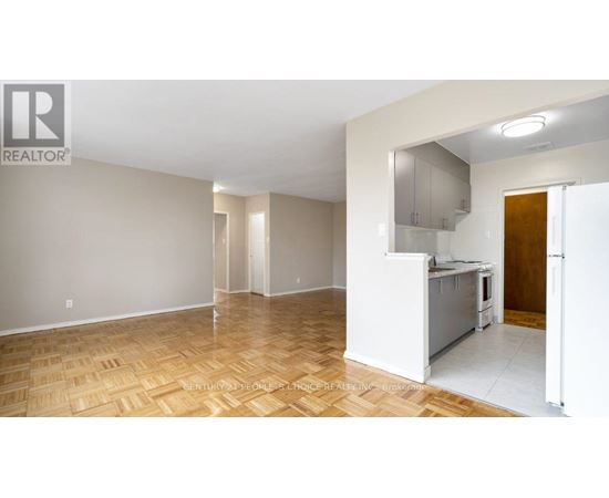 A large empty room with wooden flooring and a kitchen area in the background.