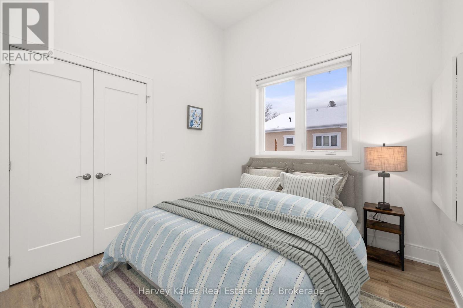 A bedroom with a bed, a nightstand with a lamp, and a window showing a view of a house.
