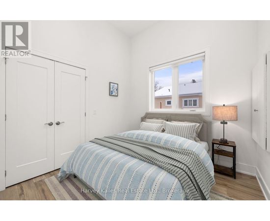 A bedroom with a bed, a nightstand with a lamp, and a window showing a view of a house.