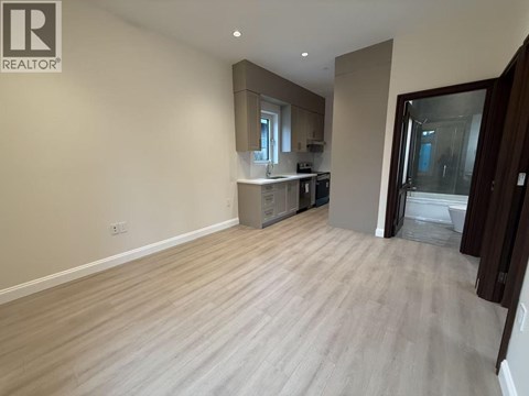 A spacious room with light wood flooring and a kitchen area in the background.