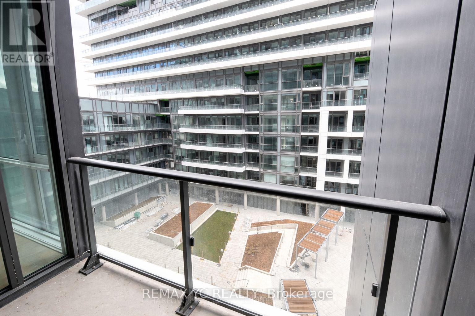 A balcony with a glass door and a metal railing overlooks a courtyard with a grassy area and several benches.