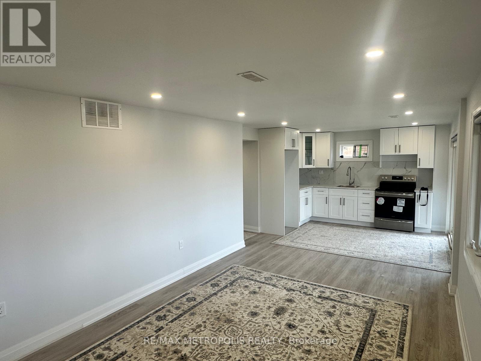 A well-lit, empty room with a rug on the floor and a kitchen area visible through the door.
