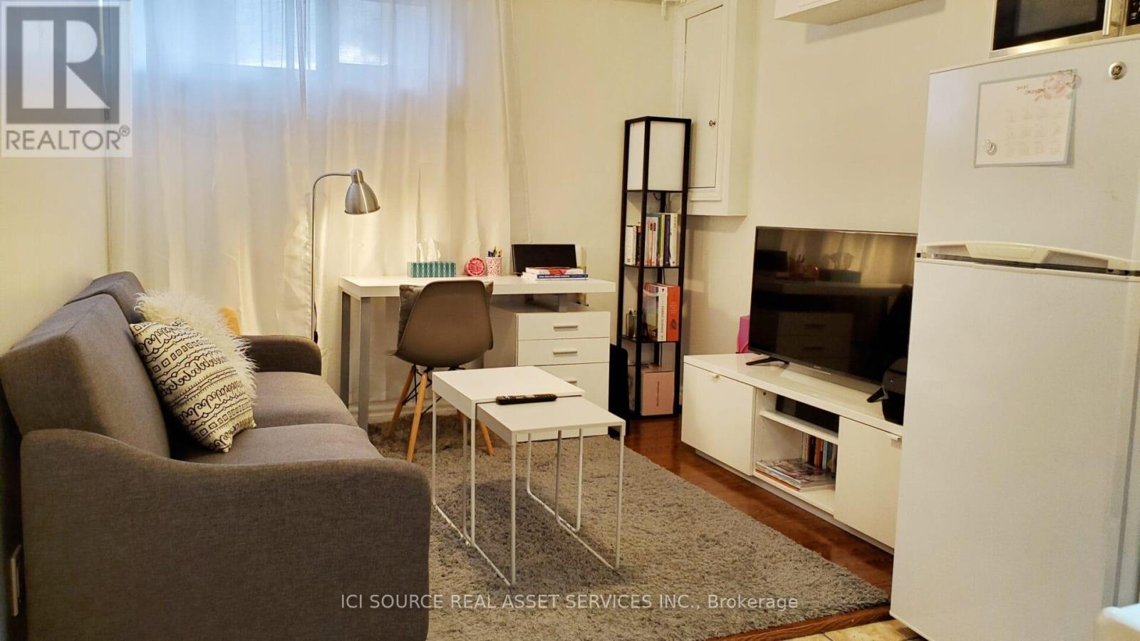 A living room with a grey couch, a white desk, and a white fridge.