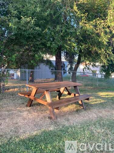 A wooden picnic table sits in a grassy area with trees and a fence in the background.