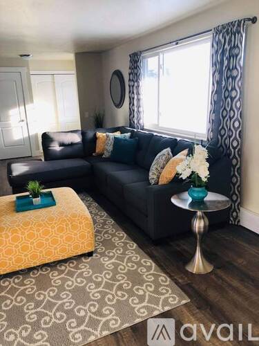 A living room with a black couch, a yellow ottoman, and a small table with a vase of flowers.