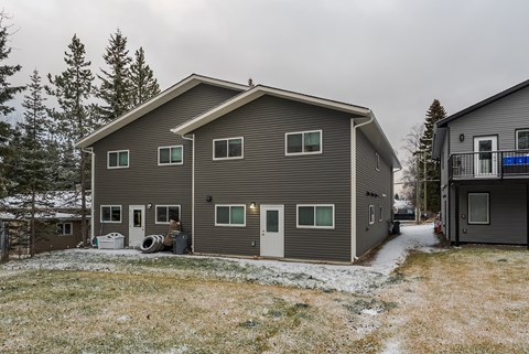 A grey house with a white door and windows is surrounded by trees and snow.
