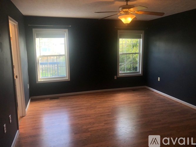 A room with dark walls and wooden flooring, featuring a ceiling fan and two windows.