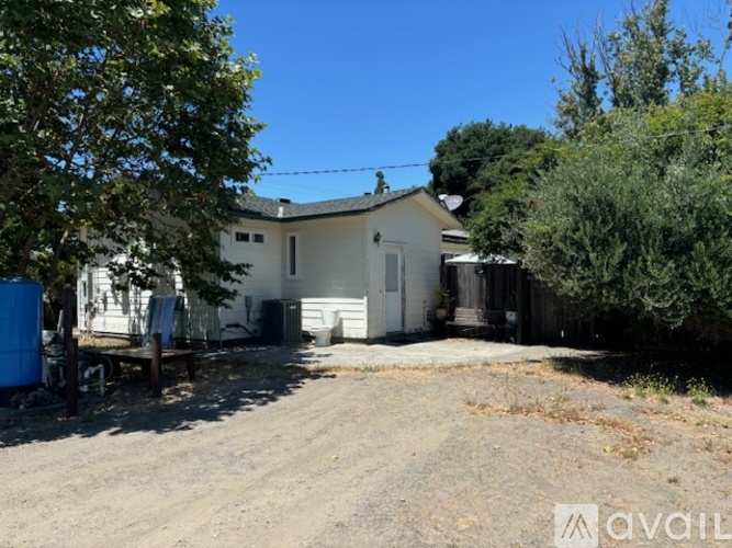 A house with a white exterior is surrounded by trees and has a dirt driveway.