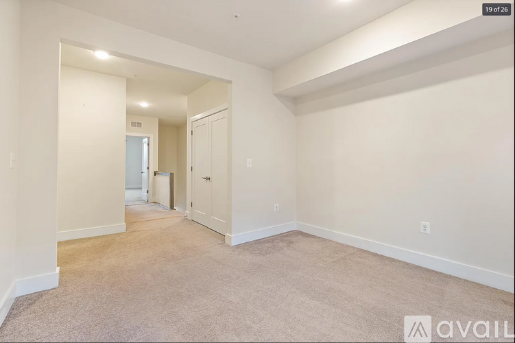 A spacious room with beige carpeting and white walls, with a door on the right and a window on the left.