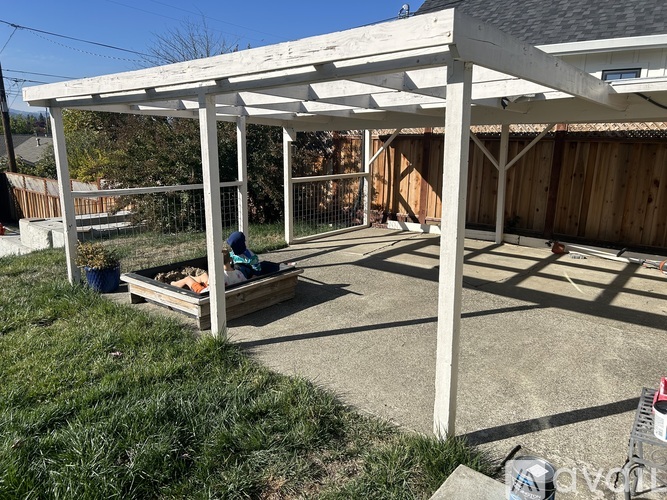 A person is sitting on a bench under a white pergola in a backyard.