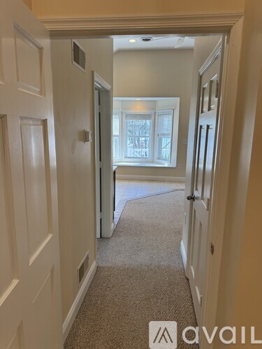 A hallway with a carpeted floor and white walls leading to a bright room with a window.