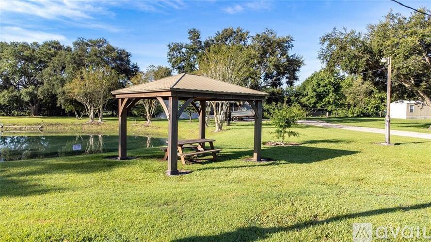 A gazebo is situated in a grassy area with a pond and trees in the background.