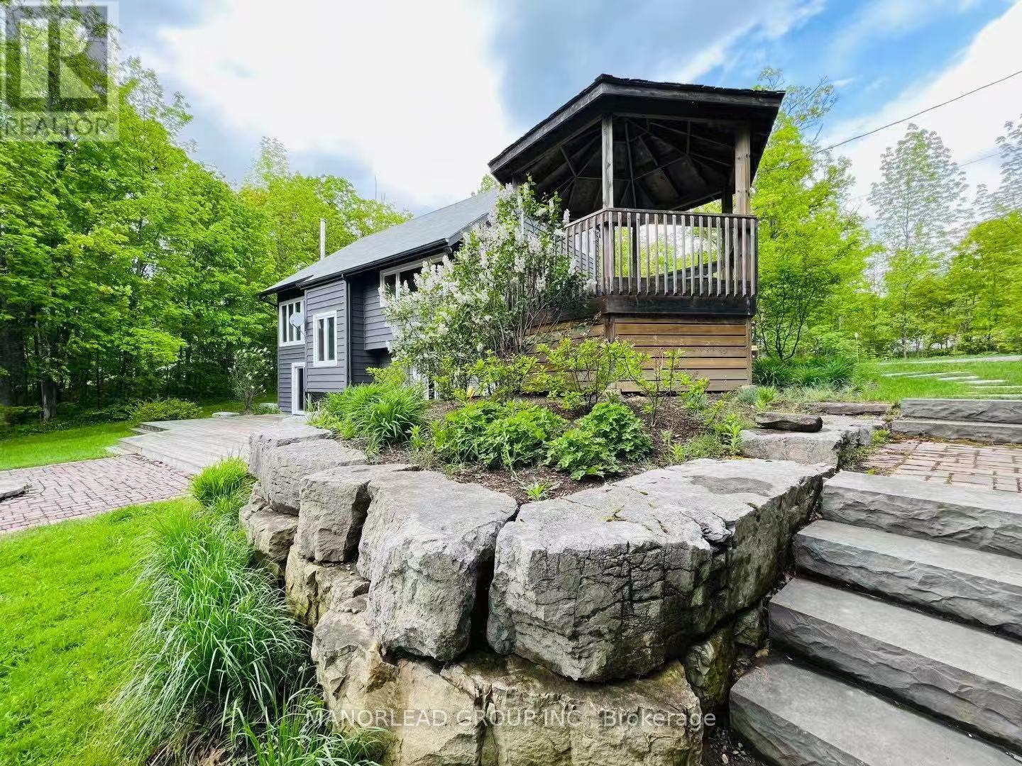 A house with a balcony is surrounded by greenery and a stone wall.