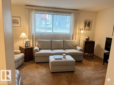 A living room with a grey sofa, a small table with a lamp and a picture frame, and a window with curtains.