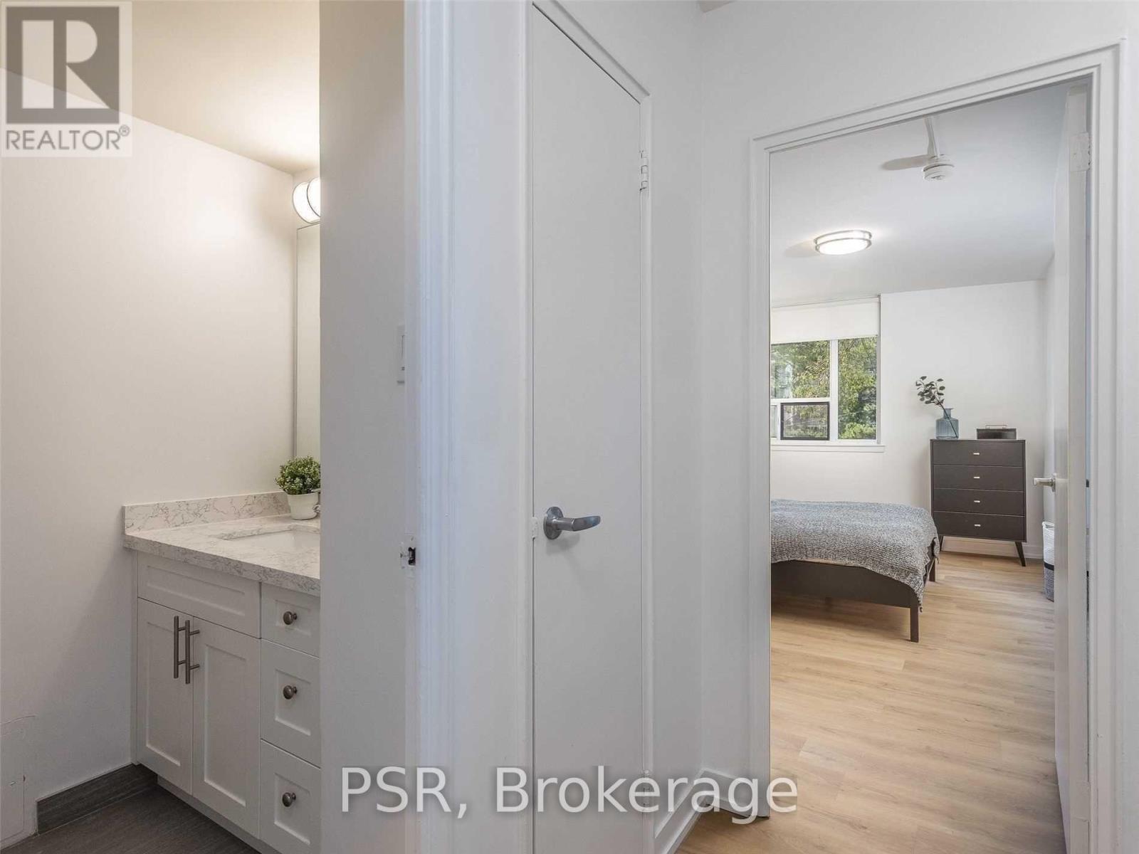 A white bathroom with a sink, mirror, and a bedroom with a bed and dresser in the background.