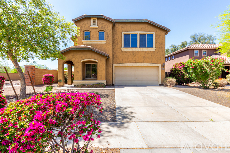 A house with a brown stucco exterior and a white garage door is surrounded by greenery and pink flowers.