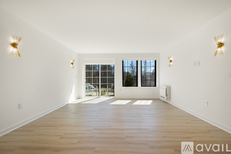 A spacious room with wooden floors and white walls, featuring a large window with a view of trees and a sliding glass door.