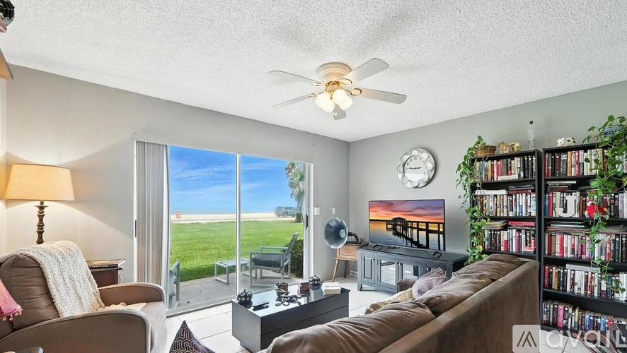 A living room with a brown couch, a fan, and a large window overlooking a green field.