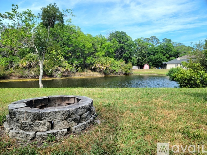A stone fire pit sits in the middle of a grassy field with a tree and a body of water in the background.