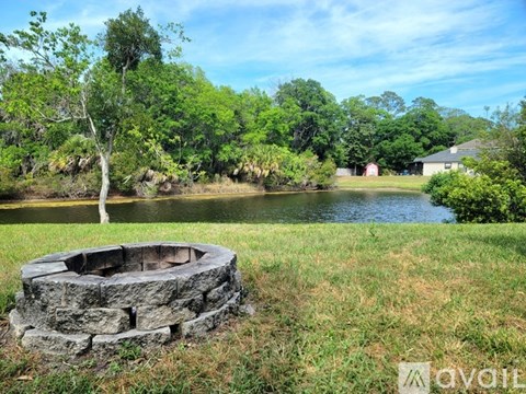 A stone fire pit sits in the middle of a grassy field with a tree and a body of water in the background.