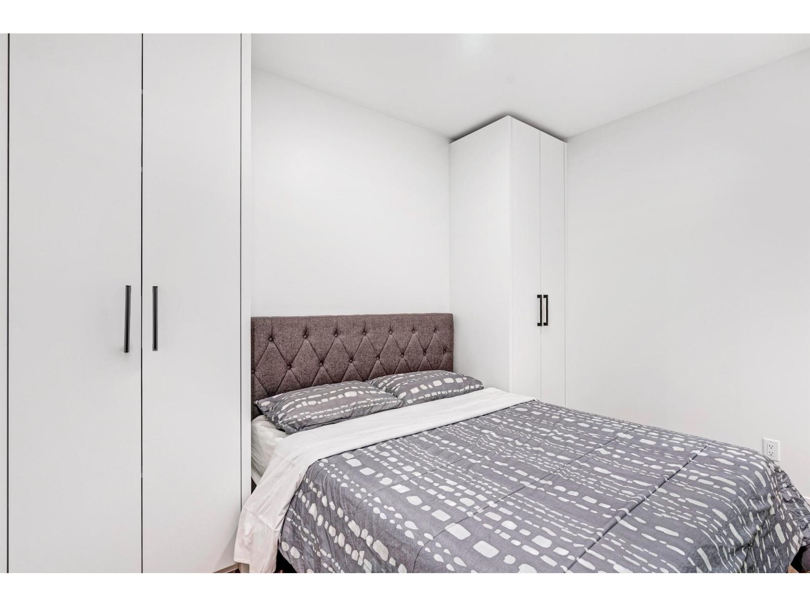 A bed with a grey patterned duvet sits in a white bedroom with a white wardrobe.