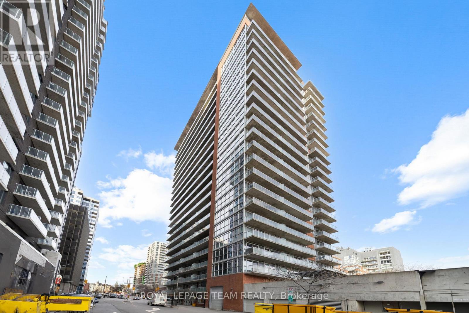 A tall building with a brown top stands in front of a smaller building with a yellow fence in the foreground.
