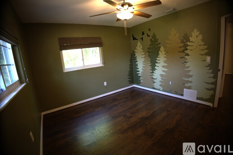 A room with a wood floor, a window with blinds, a ceiling fan, and a wall mural of trees and birds.
