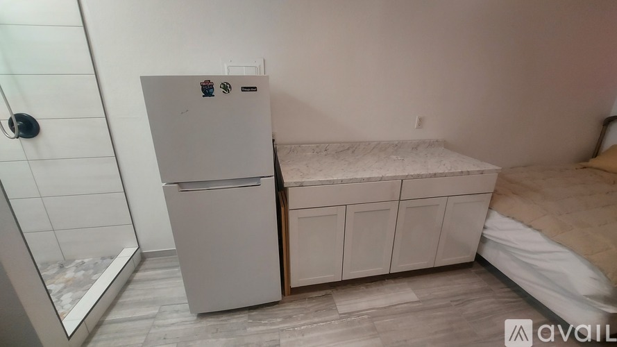 A small bedroom with a white fridge and a bed with a tan comforter.