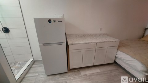 A small bedroom with a white fridge and a bed with a tan comforter.