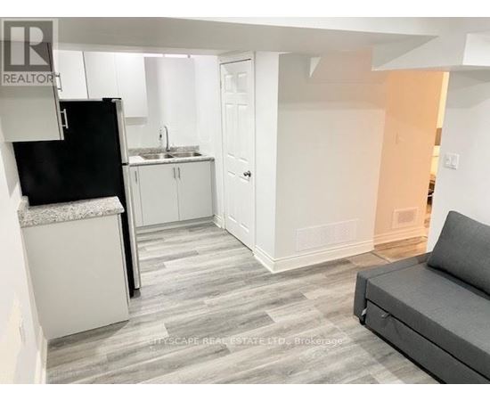A living room with a grey couch and a kitchen area with white cabinets and a black fridge.