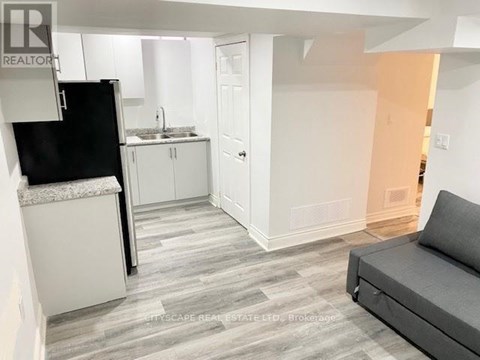 A living room with a grey couch and a kitchen area with white cabinets and a black fridge.