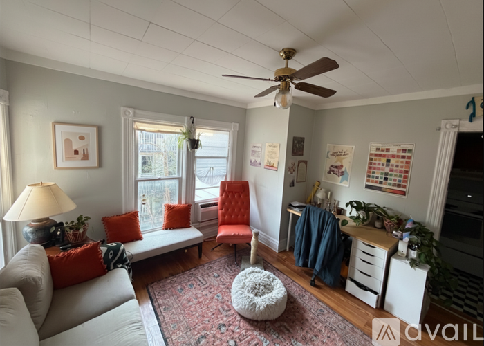 A living room with a grey couch, a red chair, a white ottoman, and a ceiling fan.