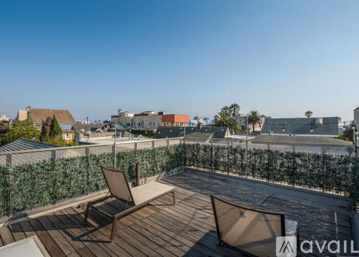 A wooden deck with two chairs and a table is overlooking a hedge and buildings.