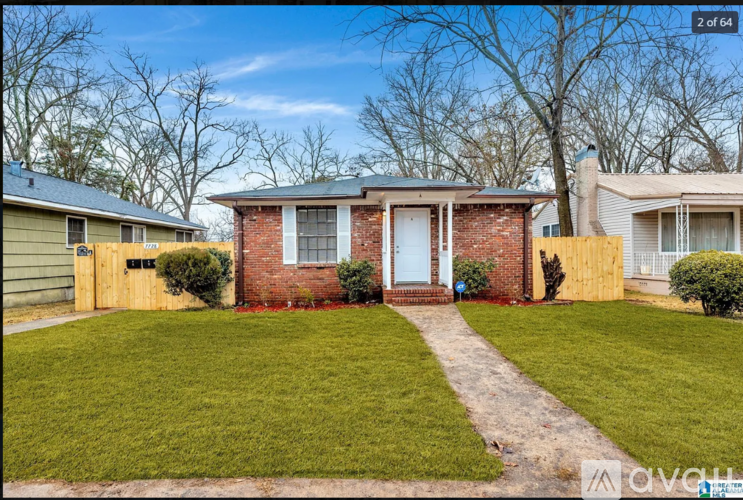 A house with a red brick facade and a white door is surrounded by a green lawn and a wooden fence.