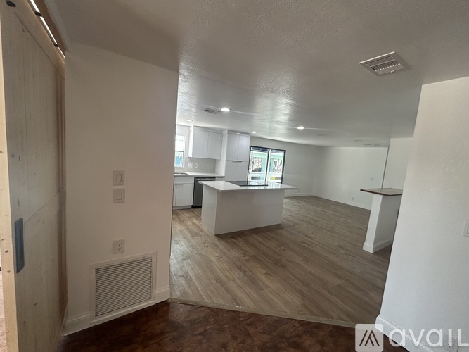 A room with a white ceiling and wooden flooring with a kitchen area in the background.