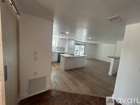 A room with a white ceiling and wooden flooring with a kitchen area in the background.