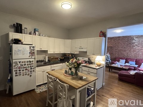 A kitchen with a refrigerator covered in magnets, a wooden table with chairs, and a brick wall in the background.