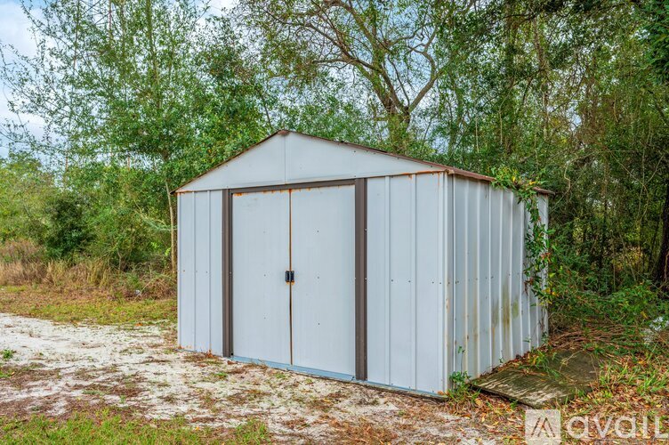 A small white metal shed with a brown door is surrounded by trees and grass.