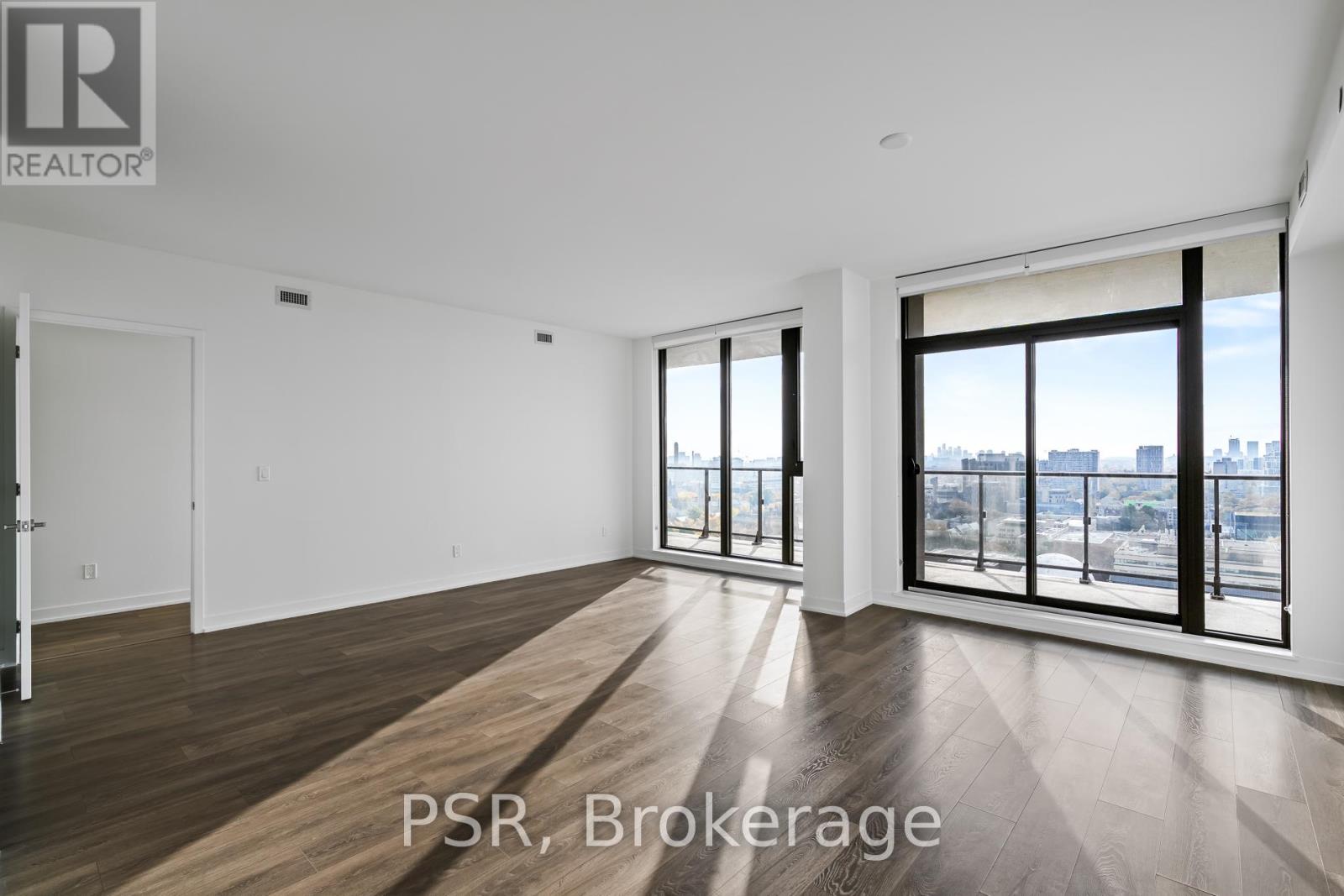 A spacious room with wooden flooring and large windows overlooking a cityscape.