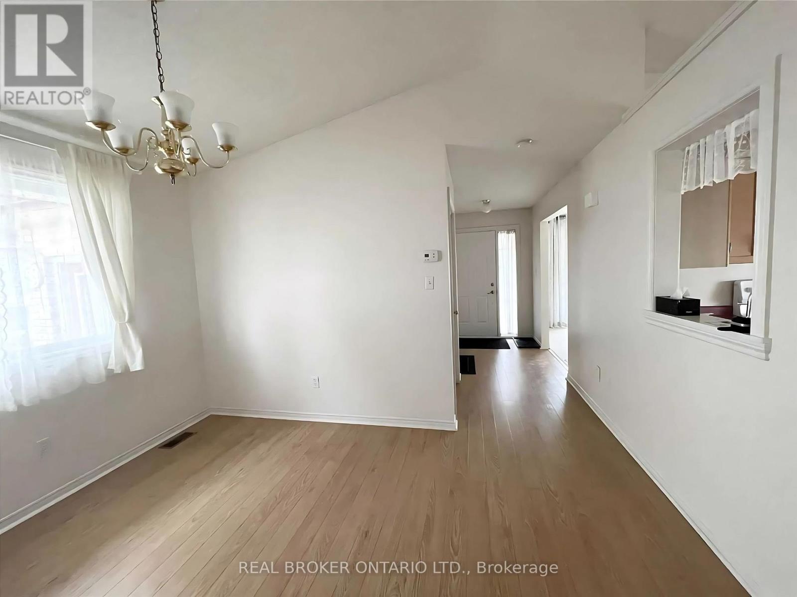 A spacious room with wooden flooring and white walls, featuring a chandelier and a window with curtains.