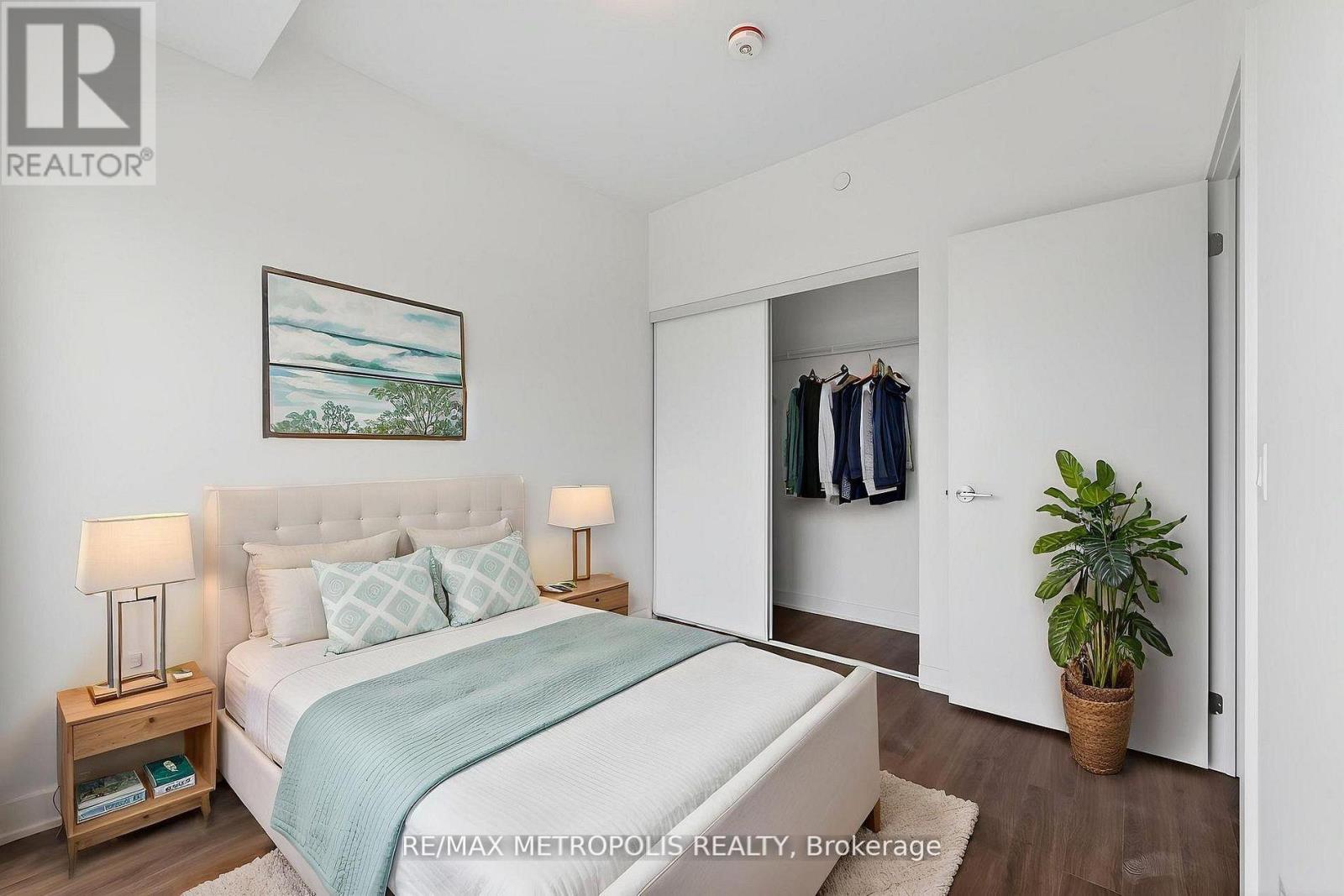 A bedroom with a bed, a nightstand with a lamp, a painting on the wall, a wardrobe with clothes hanging, and a potted plant.