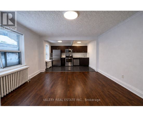 A room with wood flooring and a kitchenette in the background.
