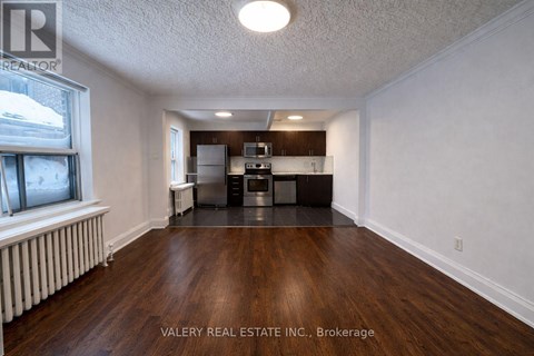 A room with wood flooring and a kitchenette in the background.